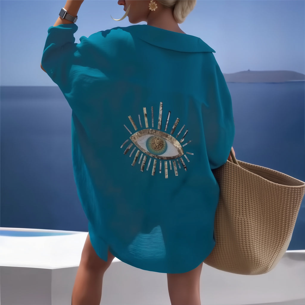 Woman wearing turquoise Costa Coral bikini cover-up with eye embroidery, holding woven beach bag by ocean.