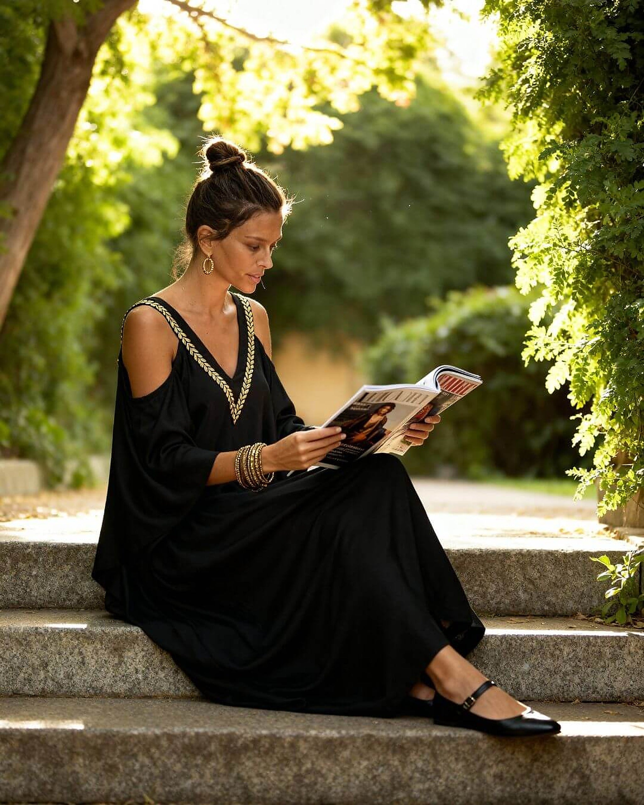 Woman wearing black cold-shoulder bikini top with gold trim details, sitting by tree-lined path reading magazine