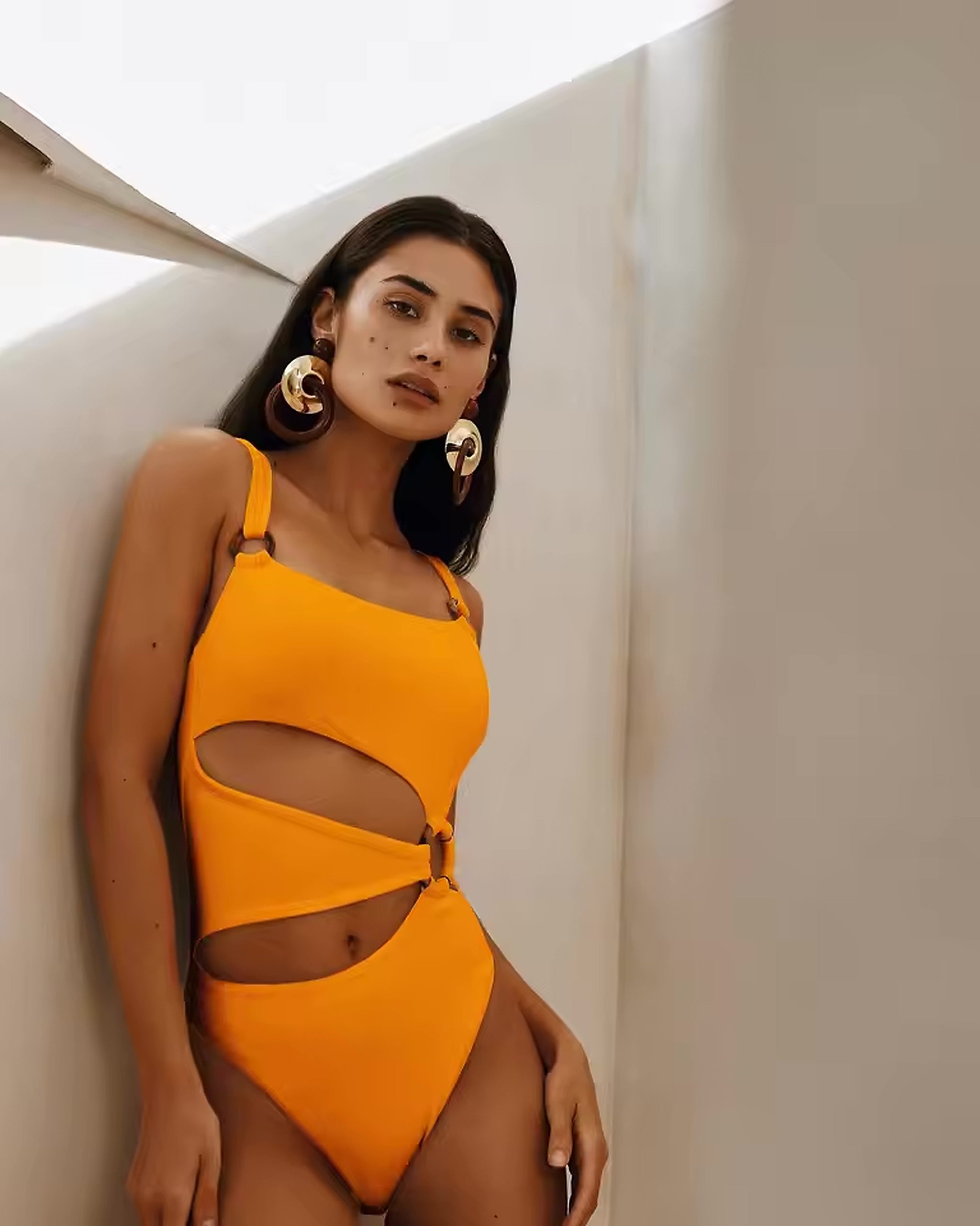 Woman wearing vibrant orange cutout bikini with adjustable straps and high-waisted bottom against white architectural