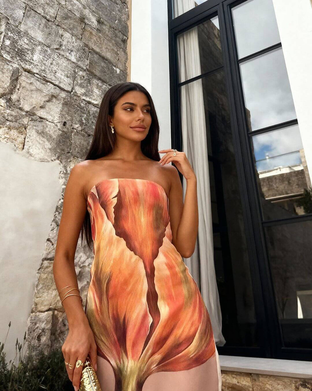 Woman wearing coral and gold gradient strapless dress by Maison Amara, posed against stone wall and black window frame.