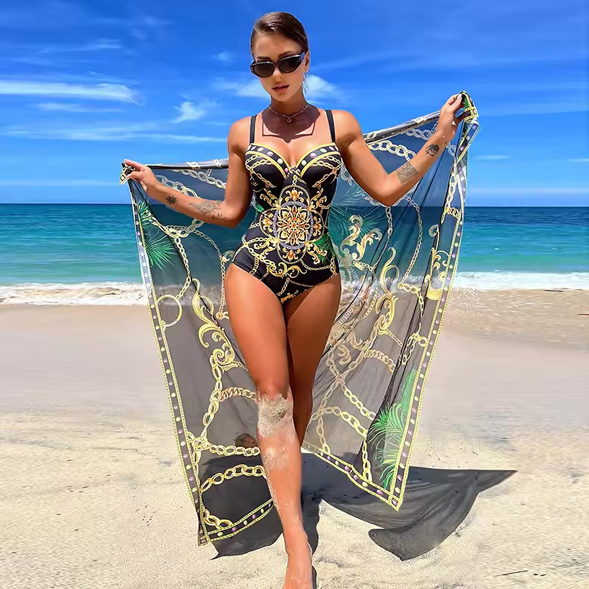 Woman in black and gold patterned one-piece swimsuit holding silk sarong on beach with ocean backdrop